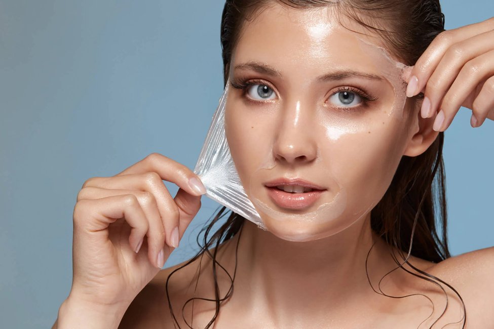 A woman peels a clear mask or layer off her face, revealing smooth skin.